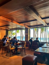 A cozy indoor seating area with wooden furniture and a large window offering a stunning view of palm trees and a beach with the ocean in the background. The warm lighting from the ceiling and the natural sunlight create a relaxing atmosphere. Several people are seated, enjoying the ambiance and the view.