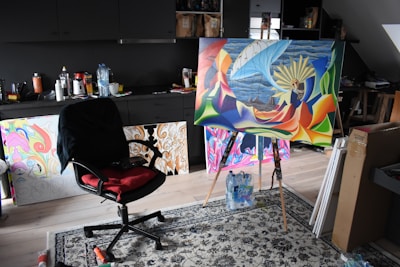 A creative studio space with a painting on an easel. The painting features bright, abstract colors and a surreal scene with a ship and large wheel. The room contains a black office chair with a red cushion, surrounded by various art supplies and multiple finished and unfinished artworks. The background has dark cabinets and some cluttered surfaces.