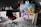 A creative studio space with a painting on an easel. The painting features bright, abstract colors and a surreal scene with a ship and large wheel. The room contains a black office chair with a red cushion, surrounded by various art supplies and multiple finished and unfinished artworks. The background has dark cabinets and some cluttered surfaces.