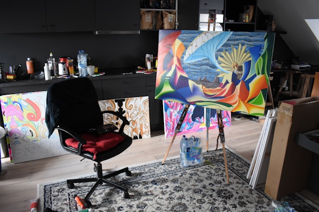 A creative studio space with a painting on an easel. The painting features bright, abstract colors and a surreal scene with a ship and large wheel. The room contains a black office chair with a red cushion, surrounded by various art supplies and multiple finished and unfinished artworks. The background has dark cabinets and some cluttered surfaces.