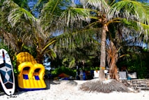 A tropical beach setting with lush palm trees providing shade. In the foreground, a yellow and blue inflatable raft and a paddleboard are leaning against the trees. The sand is white and the scene is adorned with various beach gear, suggesting a relaxed and leisurely atmosphere.