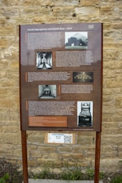An informational signboard mounted on a metal stand, set against a textured stone wall. The board is titled 'North Segregation and Death Row - 1858' and provides historical information about a prison, including images of the building, interior corridors, and an electric chair. There is text detailing the history and some notable events related to the prison experience and its segregation unit. Below the main sign, there is a QR code with a small label mentioning 'Look Inside!' and 'Move 360 Solitary.'