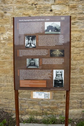 An informational signboard mounted on a metal stand, set against a textured stone wall. The board is titled 'North Segregation and Death Row - 1858' and provides historical information about a prison, including images of the building, interior corridors, and an electric chair. There is text detailing the history and some notable events related to the prison experience and its segregation unit. Below the main sign, there is a QR code with a small label mentioning 'Look Inside!' and 'Move 360 Solitary.'