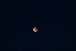 A dark night sky with a reddish-orange celestial body, likely a lunar eclipse, centered against a backdrop of stars scattered throughout the deep blue expanse.