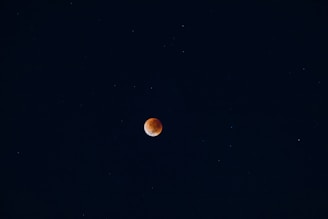 A dark night sky with a reddish-orange celestial body, likely a lunar eclipse, centered against a backdrop of stars scattered throughout the deep blue expanse.