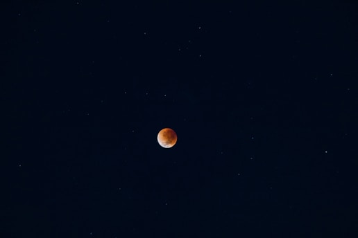 A dark night sky with a reddish-orange celestial body, likely a lunar eclipse, centered against a backdrop of stars scattered throughout the deep blue expanse.