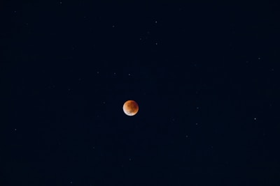 A dark night sky with a reddish-orange celestial body, likely a lunar eclipse, centered against a backdrop of stars scattered throughout the deep blue expanse.