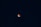 A dark night sky with a reddish-orange celestial body, likely a lunar eclipse, centered against a backdrop of stars scattered throughout the deep blue expanse.