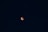 A dark night sky with a reddish-orange celestial body, likely a lunar eclipse, centered against a backdrop of stars scattered throughout the deep blue expanse.