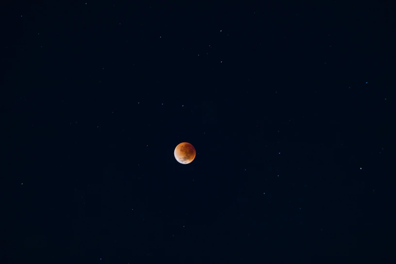 A dark night sky with a reddish-orange celestial body, likely a lunar eclipse, centered against a backdrop of stars scattered throughout the deep blue expanse.