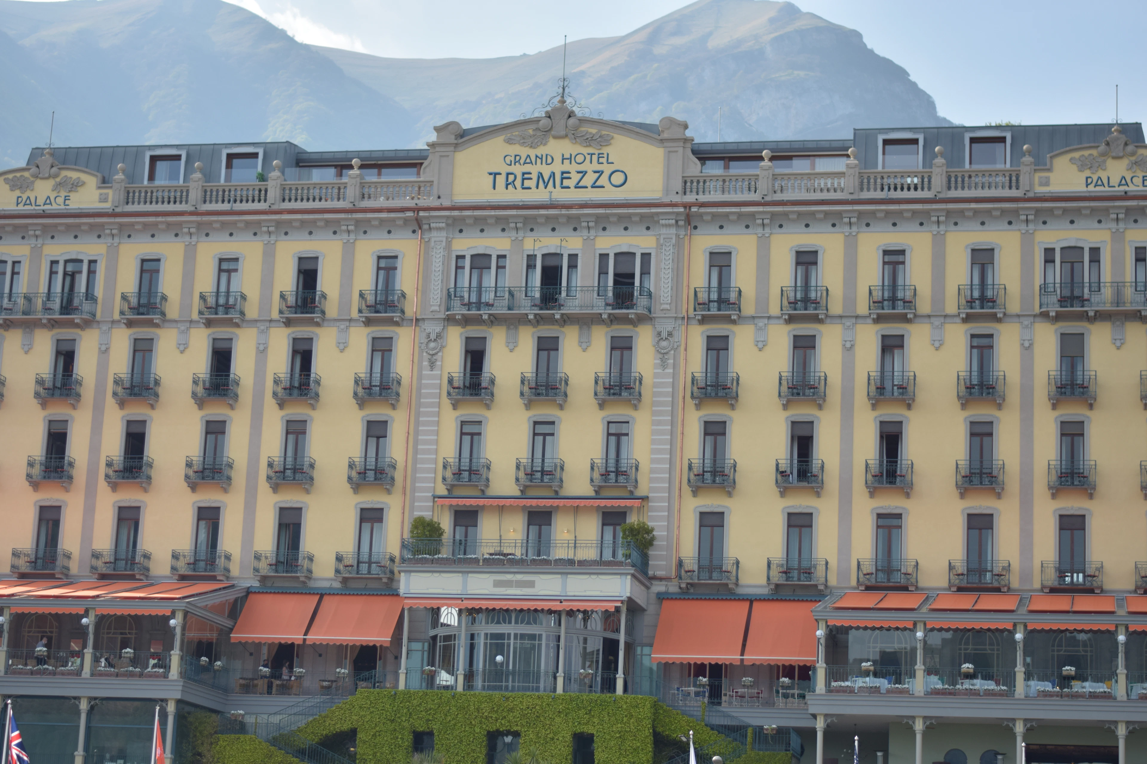 The pale yellow facade of the Grand Hotel Tremezzo on Lake Como, its name lettered across the pediment above six storeys of wrought-iron balconies, orange terrace awnings along the ground floor and a bare grey mountain rising behind the roof.