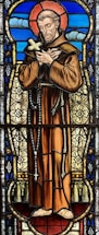 A stained glass depiction of a man with a halo, dressed in a brown robe and holding a cross, surrounded by intricate patterns and vibrant colors. The background features blue tones with cloud-like shapes.