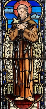 A stained glass depiction of a man with a halo, dressed in a brown robe and holding a cross, surrounded by intricate patterns and vibrant colors. The background features blue tones with cloud-like shapes.