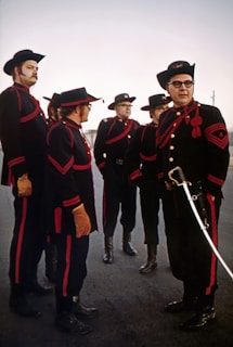 A group of men dressed in elaborate, matching uniforms consisting of dark clothes with red accents, hats, gloves, and boots. One man, standing at the forefront, holds a sword. The men display serious expressions while standing on an outdoor paved area under a clear sky.