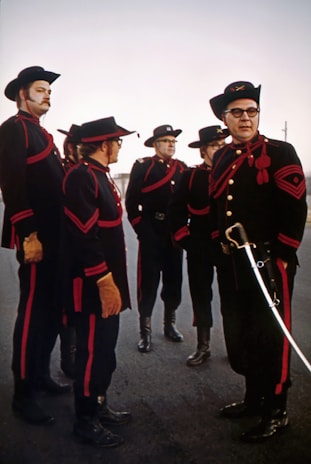 A group of men dressed in elaborate, matching uniforms consisting of dark clothes with red accents, hats, gloves, and boots. One man, standing at the forefront, holds a sword. The men display serious expressions while standing on an outdoor paved area under a clear sky.
