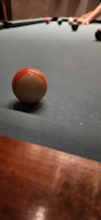 A close-up view of a pool table with several billiard balls resting on the green felt surface. A hand is visible in the background, poised to take a shot.