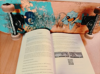 A skateboard with a colorful design and worn wheels is positioned on a wooden surface above an open book. The book contains French text, a small QR code, and illustrations of a skateboard with different foot placements labeled 'Goofy' and 'Regular.'