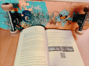 A skateboard with a colorful design and worn wheels is positioned on a wooden surface above an open book. The book contains French text, a small QR code, and illustrations of a skateboard with different foot placements labeled 'Goofy' and 'Regular.'