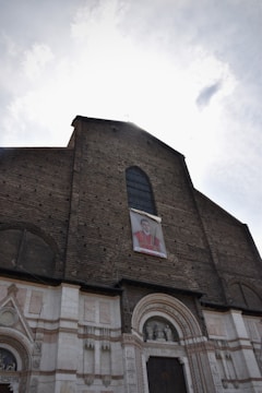 A historic brick church facade is seen with a large arched window and an entrance adorned with decorative stone carvings. A framed image is displayed prominently above the archway, featuring a person in ceremonial robes.