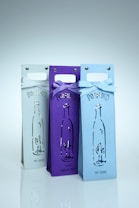 Three wine bottle bags are arranged side by side, each featuring a cutout design of a bottle. The bags display the word 'PORFIDIO' and are adorned with bows at the top, with colors ranging from white to purple to light blue.