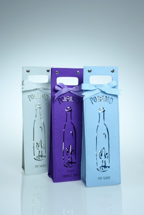Three wine bottle bags are arranged side by side, each featuring a cutout design of a bottle. The bags display the word 'PORFIDIO' and are adorned with bows at the top, with colors ranging from white to purple to light blue.