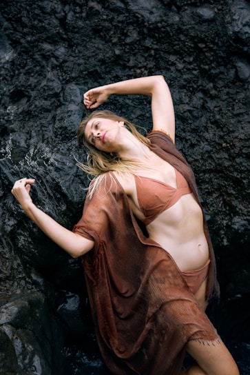 A woman in a brown bikini and cover-up poses dramatically against dark, rugged rocks. Her arms are gracefully stretched above her as she leans back, highlighting a sense of movement and elegance.