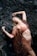 A woman in a brown bikini and cover-up poses dramatically against dark, rugged rocks. Her arms are gracefully stretched above her as she leans back, highlighting a sense of movement and elegance.
