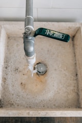A water tap with a green handle is above a stone sink, and water is flowing from the tap. The sink is filled with soapy water, and bubbles can be seen around the drain.