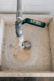 A water tap with a green handle is above a stone sink, and water is flowing from the tap. The sink is filled with soapy water, and bubbles can be seen around the drain.