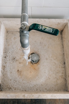 A water tap with a green handle is above a stone sink, and water is flowing from the tap. The sink is filled with soapy water, and bubbles can be seen around the drain.