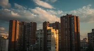 Tall residential buildings made of brick and concrete set against a backdrop of a partly cloudy blue sky. The sunlight reflects warmly off the surfaces of the buildings, casting shadows and highlighting textures.