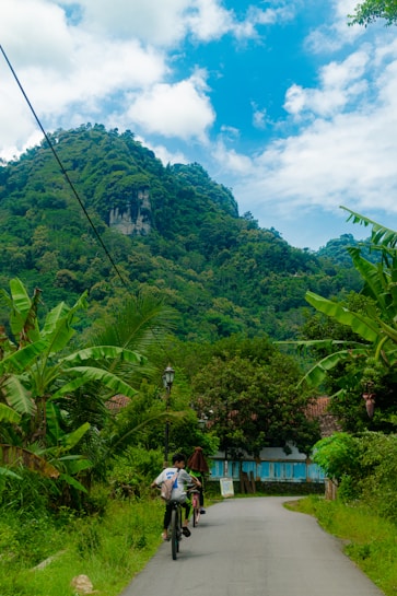 A lush, green landscape with a narrow road winding through a rural setting surrounded by dense trees and vegetation. Two people are cycling down the road toward a small, rustic building with a red roof. A steep, forest-covered hill rises majestically in the background under a vibrant blue sky with scattered white clouds.