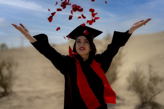 A person wearing a graduation cap and gown with a red scarf, arms raised, celebrating in a desert landscape. Red rose petals are suspended mid-air, creating a festive atmosphere against the clear blue sky.