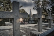 Several granite crosses and a tall obelisk monument with a red star at the top are situated in a cemetery under a grim, cloudy sky. The area is surrounded by trees and there's a paved pathway visible, along with some vegetation and fencing.