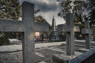 Several granite crosses and a tall obelisk monument with a red star at the top are situated in a cemetery under a grim, cloudy sky. The area is surrounded by trees and there's a paved pathway visible, along with some vegetation and fencing.