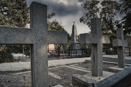 Several granite crosses and a tall obelisk monument with a red star at the top are situated in a cemetery under a grim, cloudy sky. The area is surrounded by trees and there's a paved pathway visible, along with some vegetation and fencing.