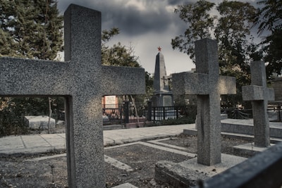 Several granite crosses and a tall obelisk monument with a red star at the top are situated in a cemetery under a grim, cloudy sky. The area is surrounded by trees and there's a paved pathway visible, along with some vegetation and fencing.