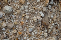 A close-up view of a rough, rocky surface with various sizes of stones and gravel. The stones are unevenly distributed with a mix of grey, brown, and reddish hues, giving the surface a textured and earthy appearance.