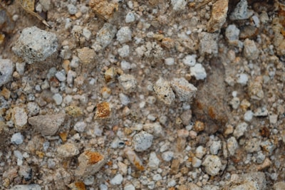 A close-up view of a rough, rocky surface with various sizes of stones and gravel. The stones are unevenly distributed with a mix of grey, brown, and reddish hues, giving the surface a textured and earthy appearance.