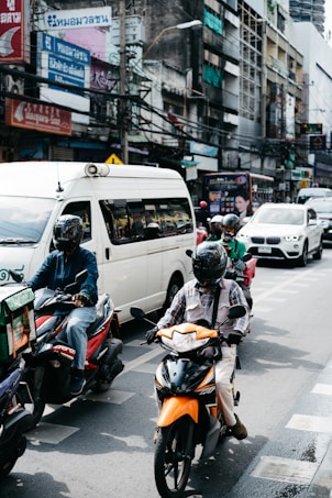 Motorcycles and a van are navigating through a busy city street lined with buildings and various signs in a foreign language. Several riders are wearing helmets, and some vehicles are visible in the background amid a bustling urban environment.