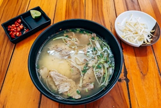 A black bowl filled with a clear broth containing sliced meat, green herbs, and vegetables is placed on a wooden table. On the side, there is a small rectangular dish with sliced red chilies and a lime wedge. A separate white bowl contains bean sprouts.