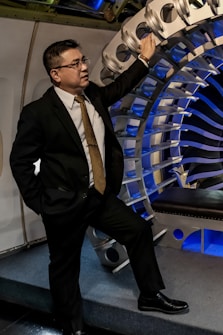 A man in a suit and tie is posing next to a large industrial turbine or engine component. The setting appears professional and technical, with a focus on machinery.