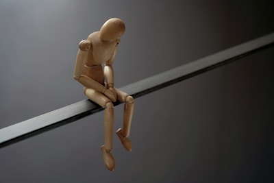 A wooden artist's mannequin sitting on a thin, horizontal beam. The figure's posture suggests introspection or contemplation, as its head is bowed and arms rest on its knees. The background is a muted grey, enhancing the minimalist and somber visual effect.