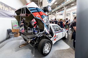 A sports car with its engine cover lifted, showcasing intricate mechanical components. The vehicle has a distinct racing livery with 'Martini' branding and is displayed in an exhibition hall filled with people. In the background, some visitors observe and discuss the exhibits.