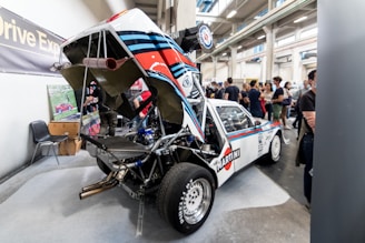 A sports car with its engine cover lifted, showcasing intricate mechanical components. The vehicle has a distinct racing livery with 'Martini' branding and is displayed in an exhibition hall filled with people. In the background, some visitors observe and discuss the exhibits.