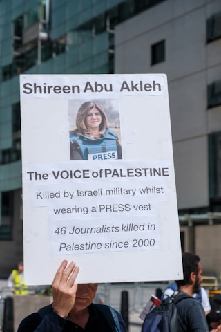 A large sign is being held in an urban environment. The sign features a photograph of a person wearing a blue press vest. Above the image, the name 'Shireen Abu Akleh' is printed. The sign also contains text that reads 'The VOICE of PALESTINE,' and mentions that the person was killed by the Israeli military while wearing a press vest. Additional text states that 46 journalists have been killed in Palestine since 2000.