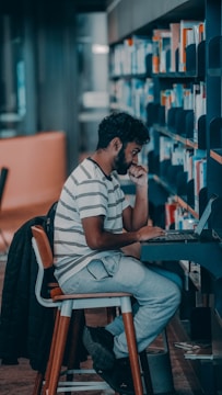 A person in a striped shirt is seated on a wooden stool, intently using a laptop in a library setting. The person is surrounded by shelves filled with books, and appears to be deep in thought, possibly researching or studying.
