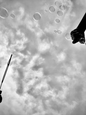 Bubbles float against a cloudy sky, with two silhouetted hands creating them using bubble wands.