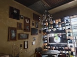 A cozy interior space features eclectic wall art and various vintage cameras displayed on shelves. The wall is adorned with an assortment of framed pictures, some depicting abstract and monochrome images. A chandelier hangs from the ceiling, adding a touch of elegance. A small, round wooden table is surrounded by chairs, creating a warm atmosphere.