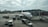 A large passenger airplane is parked at an airport gate, connected to a jet bridge. Several ground service vehicles and workers are visible, performing various operations around the aircraft. In the background, another airplane is visible on the runway, with a backdrop of distant hills and buildings under a cloudy sky.